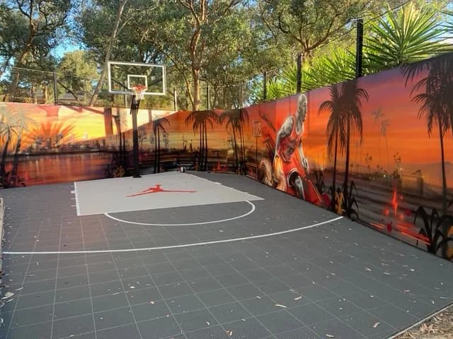 Backyard basketball half court on grey modular tiles with a hoop and a painted mural wall
